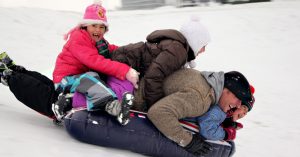 A dad having fun with his kids sliding down a snowy hill on an inner-tube.